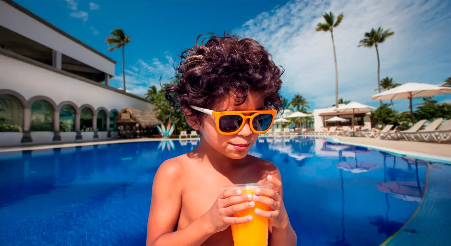 Niño con gafas naranjas tomando jugo frente a alberca en resort tropical.
